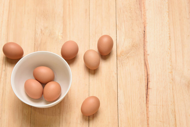 Три яйца в белой чашке и одно яйцо, помещенное вокруг чашки на светло-коричневом деревянном фоне, Вид сверху с пространством для копирования
 - Фото, изображение