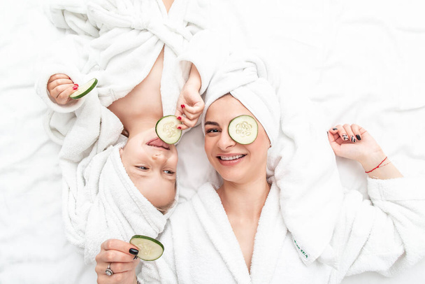 Mère et fille positives faisant masque facial appliquant des morceaux de concombre à leurs yeux, enveloppés dans une serviette de bain blanche. - Photo, image