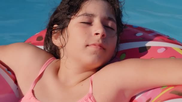11-12歳の幸せなかわいい白人の女の子は庭のスイミングプールで膨脹可能なドーナツサークルに横たわっています。子供や子供のための涼しい夏の休日。水の上のゲーム。ピンクのドーナツ。4kスローモーション - 映像、動画