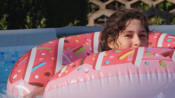 11-12 jaar oud gelukkig schattig blank meisje liggend op een opblaasbare donut cirkel in het zwembad in een tuin. Leuke zomervakantie voor kinderen en kinderen. spelletjes op het water. Roze donat. 4k slow motion - Video