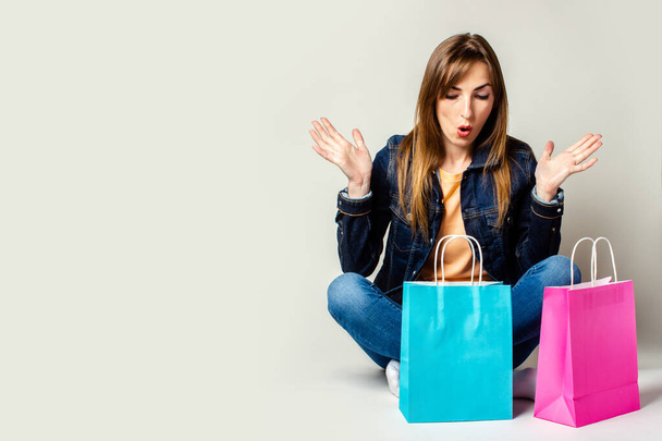 Junge Frau mit überraschtem Gesicht in Jeansjacke hält Einkaufstüten in der Hand, während sie auf dem Boden vor hellem Hintergrund sitzt. Banner. Konzepteinkauf, Verkauf, Rabatte. - Foto, Bild