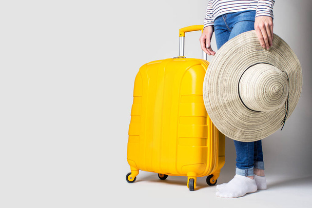 Woman holds a plastic yellow suitcase and a hat from the sun on a light background. Travel concept, flight expectation, vacation. Only legs are visible. Banner. - Фото, изображение