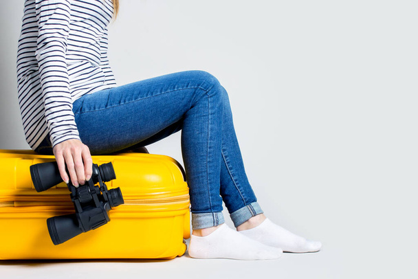 Vrouw houdt een verrekijker vast en zit op een gele plastic koffer op een lichte achtergrond. Reisconcept, vluchtverwachting, vakantie. Alleen benen zijn zichtbaar. Banner. - Foto, afbeelding