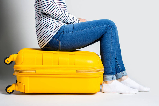 Kobieta siedzi na żółtej plastikowej walizce na jasnym tle. Koncepcja podróży, oczekiwanie na lot, wakacje. Widać tylko nogi. Sztandar. - Zdjęcie, obraz