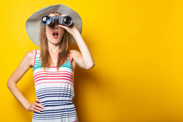 Young woman in shock in surprise with a hat and a striped dress looks in surprise with binoculars on a yellow background. - 写真・画像