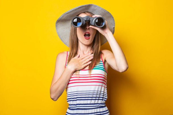 Young woman in shock in surprise with a hat and a striped dress looks in surprise with binoculars on a yellow background. - Foto, Imagem