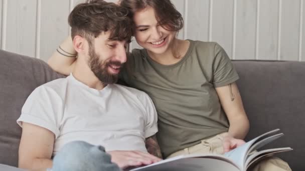 A romantic caucasian cute couple are smiling and reading a book together while sitting in the living room at home - Séquence, vidéo