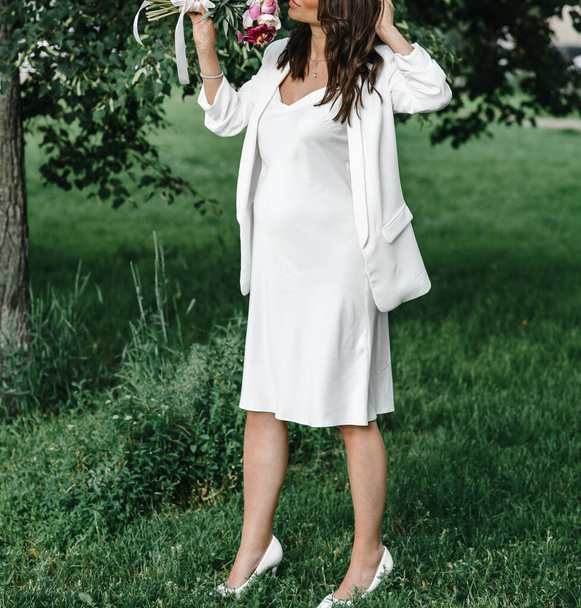 novia embarazada en un vestido blanco delgado en tirantes delgados con hombros desnudos con un ramo de peonías. maternidad
 - Foto, Imagen