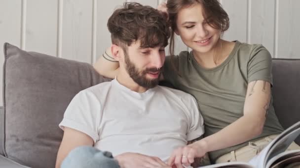 Un couple heureux sourit et lit un livre ensemble tout en étant assis dans le salon à la maison - Séquence, vidéo