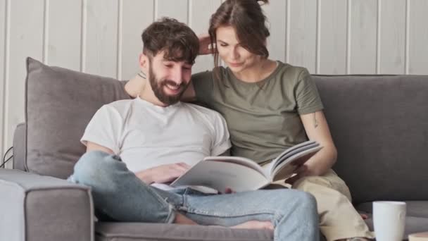 A smiling couple man and woman are reading a book together while sitting in the living room at home - Video, Çekim