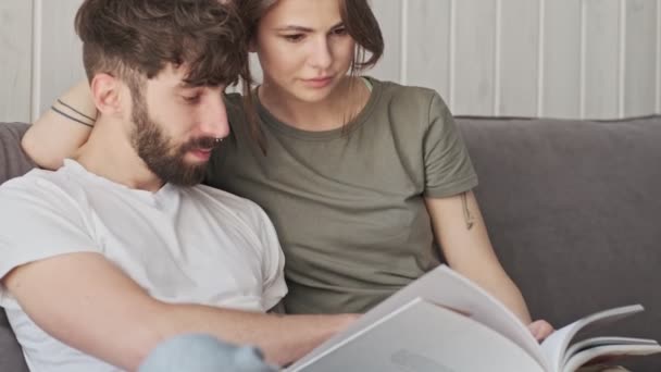 An attractive young couple man and woman are reading a book together while sitting in the living room at home - Filmagem, Vídeo