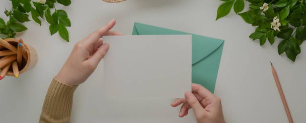 Overhead shot of female hands holding mock-up greeting card above turquoise envelope on white desk with decorations - Fotó, kép