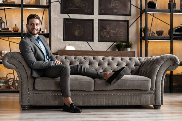 Cool and handsome. Good looking young man in full suit sitting on the sofa while spending time indoors. - Foto, Imagem