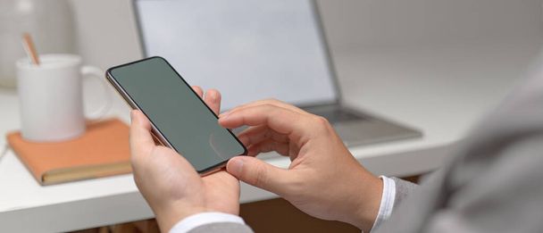 Visão de perto de mãos empreendedoras masculinas usando o smartphone mock-up enquanto se senta na mesa de escritório branca com material de escritório
  - Foto, Imagem