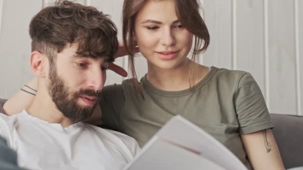 A close-up cropped view of a good-looking young couple man and woman are reading a book together while sitting in the living room at home - 映像、動画
