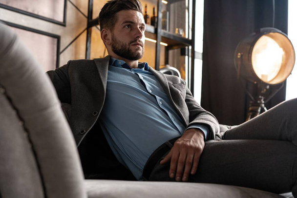 Cool and handsome. Good looking young man in full suit sitting on the sofa while spending time indoors. - Fotografie, Obrázek