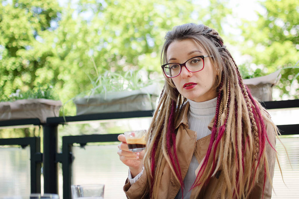 Junge Frau mit Brille, lässigem nackten Trench und langen Dreadlocks trinkt Kaffee in Outdoor-Café. - Foto, Bild