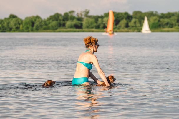 Eine ältere Frau, die mit ihrem süßen Hund Dackel im Flussschwimmen Ball spielt. Konzept Haustiere, Liebe zu Tieren. Frau im Badeanzug warf Ball nach Hundebegleiterin. - Foto, Bild