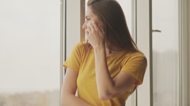 Μια όμορφη νεαρή γυναίκα κοιτάζει έξω από το παράθυρο στέκεται στο σαλόνι στο σπίτι - Πλάνα, βίντεο