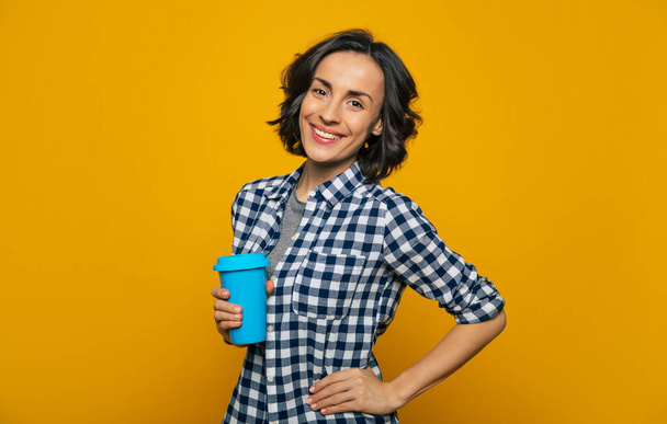 My new thermal mug! A half-length photo of a young attractive student dressed casually smiling at the camera, holding her favorite comfy blue thermal mug in her one hand, and having another on her hip - Foto, Bild
