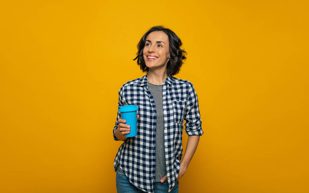 Relaxed day. Half-length photo of a relaxed  smiley girl in a checkered shirt, holding a blue cup in her right hand, looking aside and up, standing with her left hand in a pocket. - Foto, Bild