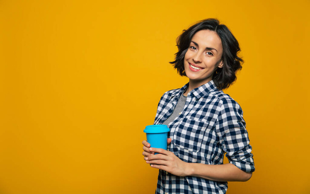 I love this day. Close-up photo of a relaxed pretty girl with bright eyes in a black and white checkered shirt, holding a blue cup in her right hand, standing aside. - Fotografie, Obrázek