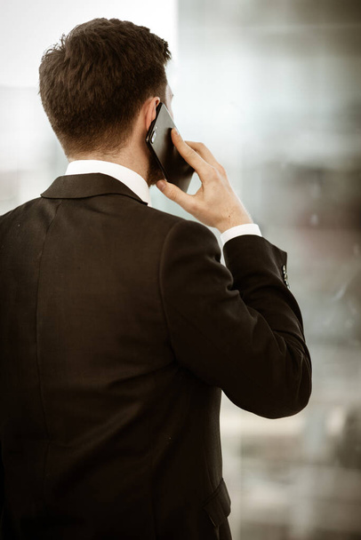 Business concept. Young businessman at the office standing and busy talking on a phone showing with hand he is resolving a very serious work problem. Man in suit indoors on glass window background - Photo, Image