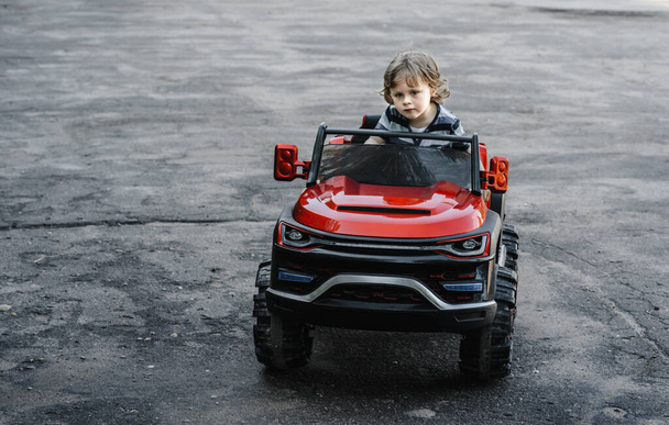 Çizgili tişörtlü kıvırcık saçlı çocuk asfalt yolda kırmızı büyük bir oyuncak araba sürüyor. Tatil günü, açık hava eğlencesi. - Fotoğraf, Görsel