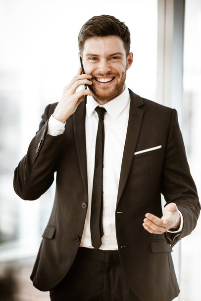 Bedrijfsconcept. Blij lachende jonge zakenman die in het kantoor staat te praten op een mobiele telefoon krijgt goed nieuws over zijn werk. Man in pak binnenshuis op glazen raamachtergrond - Foto, afbeelding