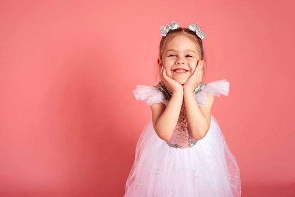 Portrait of cute little girl wearing white-pink dress, smiling and propping up her head with hands over pink background - Photo, Image