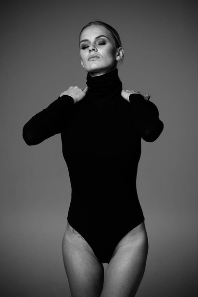 ファッションとランジェリーのコンセプト-グレーの背景にセクシーな黒のタートルネックボディコルセットの美しいブロンドの女性の肖像画。下着姿の女がスタジオライトでポーズ。白黒モノクロームで - 写真・画像
