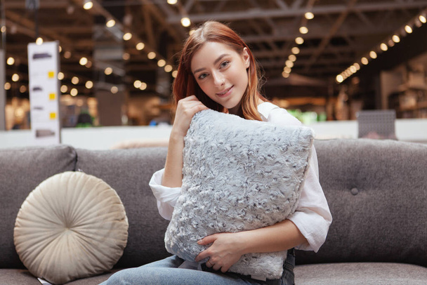 Krásná veselá žena objímající polštář, sedící na nové pohovce v obchodě s nábytkem - Fotografie, Obrázek