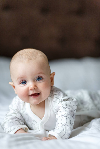 A cute little baby is smiling lying on the bed on a white sheet in the parents bedroom. Closeup portrait of newborn baby looking at camera. vertical photo - Zdjęcie, obraz