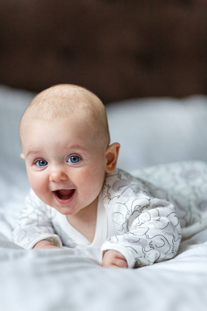A cute little baby is smiling lying on the bed on a white sheet in the parents bedroom. Closeup portrait of newborn baby looking at camera - Zdjęcie, obraz