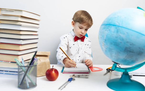 Preschool education: a little boy in a shirt and a red bow tie sits at a desk and takes notes with a pencil in a notebook. On the table are books and a globe. Portrait. - 写真・画像