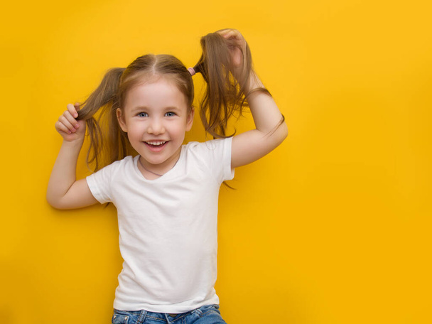 A naughty little girl in a white T-shirt is holding herself by the hair on a yellow background. Mocap. Copyspace - Фото, изображение