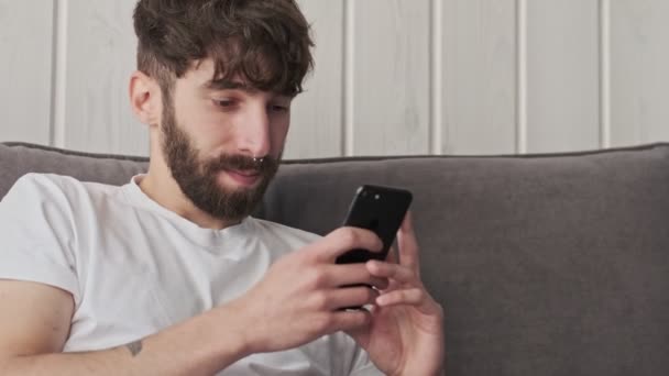 A handsome young hipster man in casual clothes is using his smartphone sitting in the living room at home - Metraje, vídeo