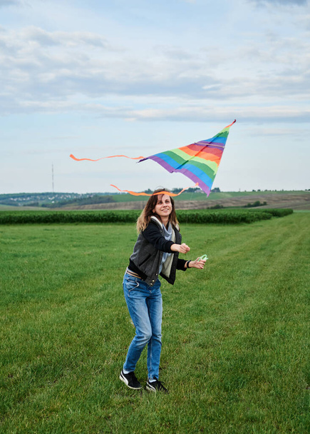 Young brunette woman, wearing casual clothes green t-shirt, playing with colorful kite on green field meadow in summer, running, jumping. Family leisure activity at natural rural landscape. - Foto, Bild