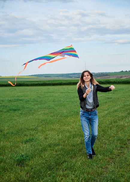 Young brunette woman, wearing casual clothes green t-shirt, playing with colorful kite on green field meadow in summer, running, jumping. Family leisure activity at natural rural landscape. - Foto, imagen