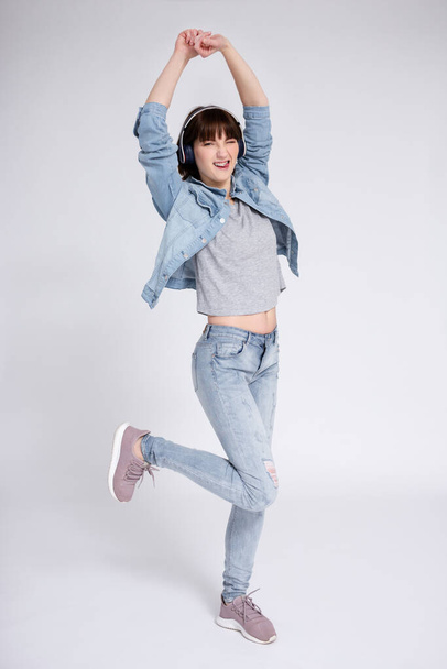 full length portrait of cheerful teenage girl with braces on teeth listening music in headphones and dancing or celebrating something over gray background - Photo, Image