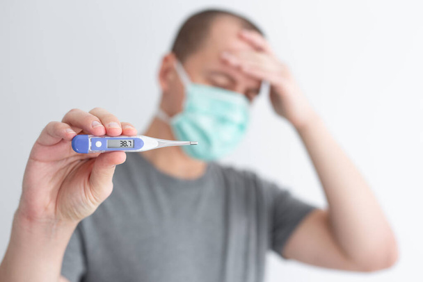 Gesundheitsfürsorge, Krankheit und Pandemiekonzept - Kranker mit Grippe oder Coronavirus und Kopfschmerzen trägt Schutzmaske und zeigt elektronisches Thermometer - Foto, Bild
