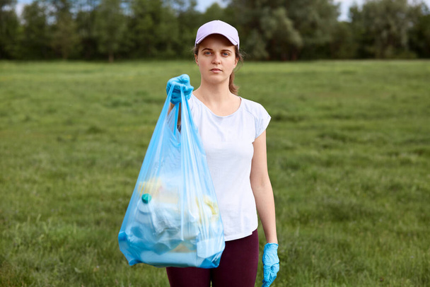 Serious lady in baseball cap holding garbage bag full of trash in hands, showing it to people, calls on to clean up our planet, fights with pollution, posing in open air. - 写真・画像