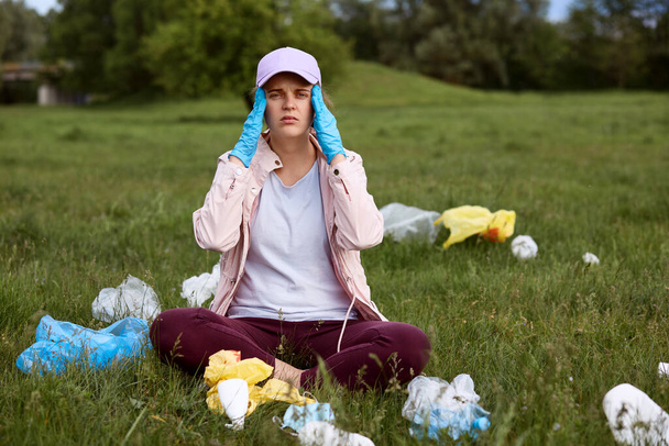 Tired environmental activist sitting on green grass with fingers on temples, exhausted female picking up trash in park, being in despair, calls for sorting litter and throw garbage in designated areas - Photo, image