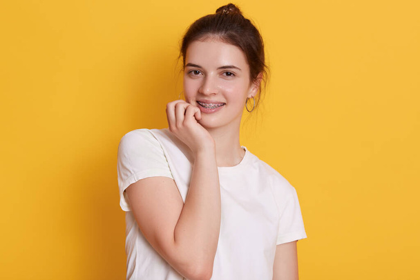 Adorable dark haired woman in brackets looking at camera with charming smile, holding her fist under chi, girl with hair bun dresses white casual t shirt. - Foto, immagini