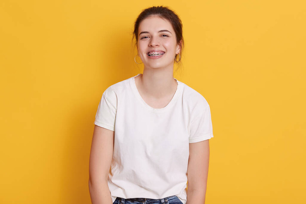 Happy smiling brunette girl with hair bun dresses white casual t shirt, posing isolated over yellow background, expressing positive emotions, looks at camera. - Фото, изображение