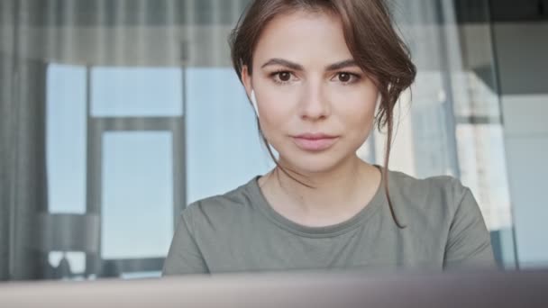 An attractive young woman with earbuds is working while using her laptop computer sitting at home - Metraje, vídeo