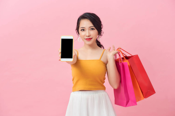 Portrét docela vzrušená dívka s barevnými nákupními taškami ukazující prázdné obrazovky mobilní telefon přes růžové pozadí. - Fotografie, Obrázek
