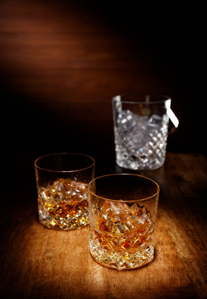 Reflektor na dwóch kryształowej szklance szkockiej whisky z wiadrem lodu w backghround, strzał na drewnianym blat stołu. - Zdjęcie, obraz