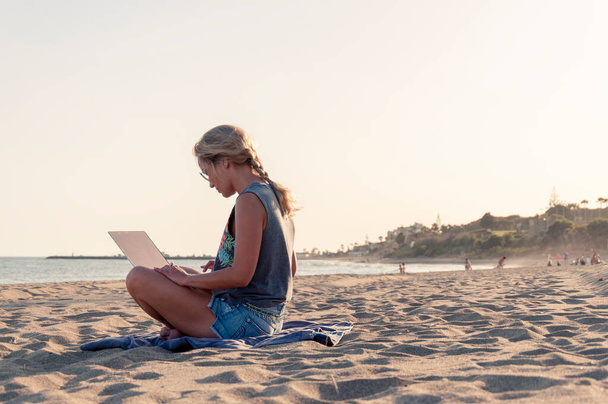 Una ragazza in pantaloncini e una t-shirt è seduta su una spiaggia sabbiosa con una vista laterale del computer portatile, sullo sfondo le persone stanno nuotando nel mare e c'è un villaggio turistico - Foto, immagini
