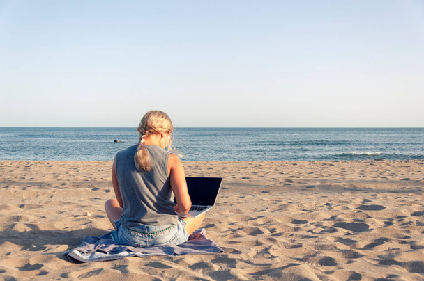 A girl with blond hair sits on a sandy beach against the  of the sea in shorts and a T-shirt and with a laptop, view from the back  - Foto, Imagem